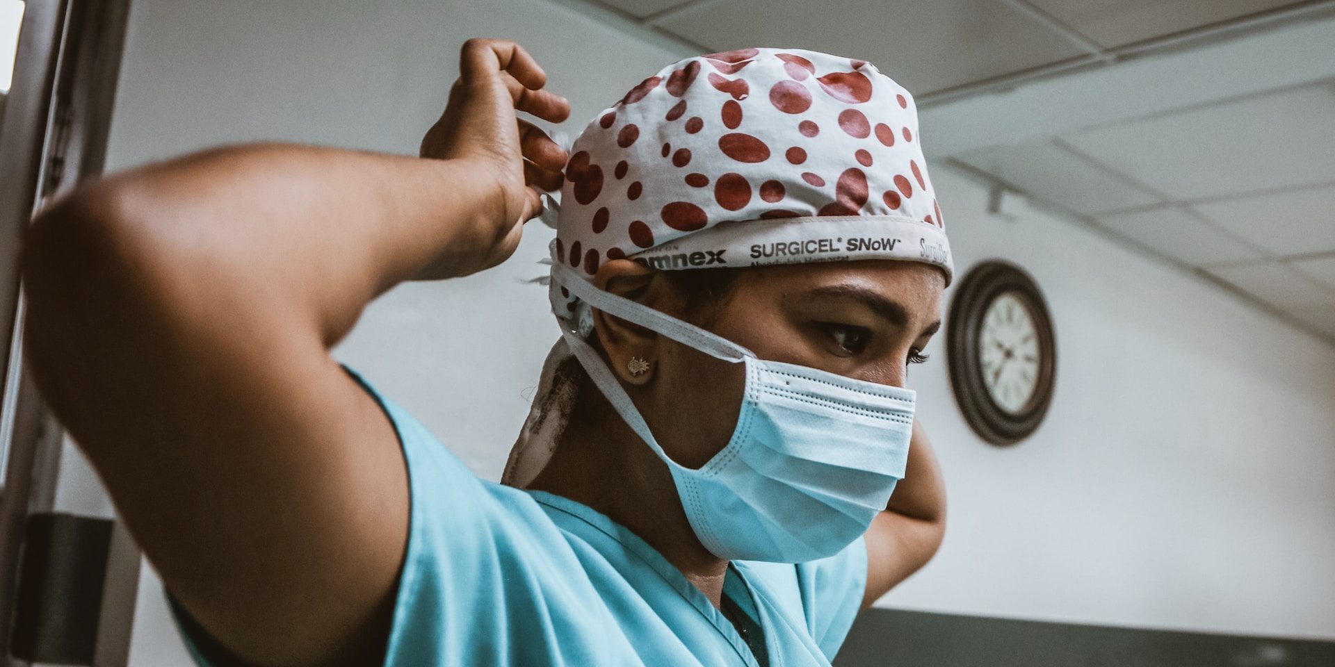 A surgeon wearing medical scrubs secures a face mask around the back of her own head.