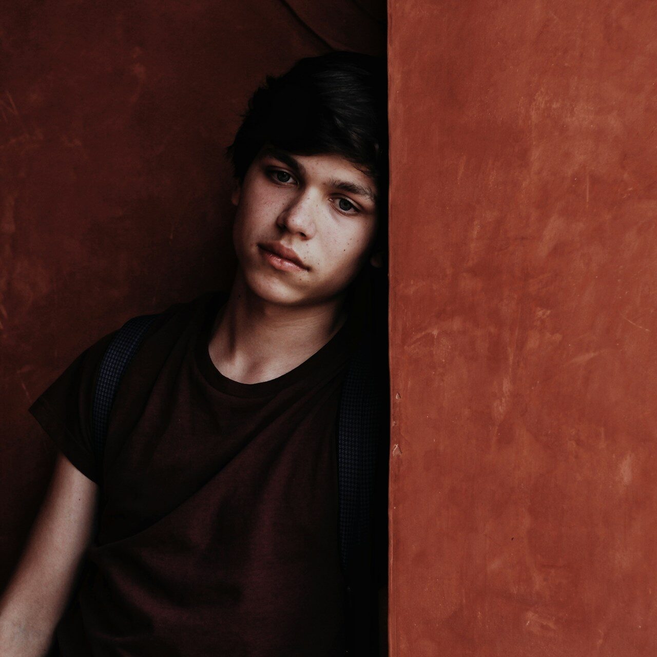 A boy stares blankly as he leans into the corner of a concrete pillar, partially obscuring him.