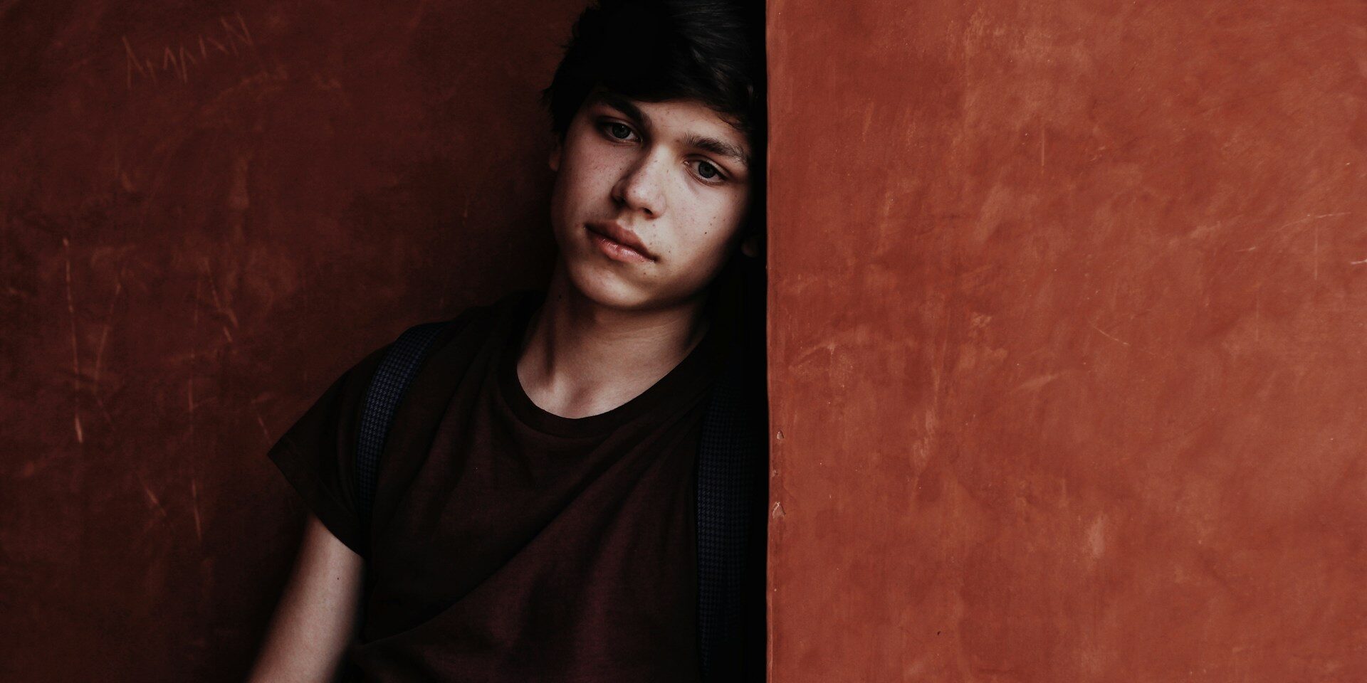 A boy stares blankly as he leans into the corner of a concrete pillar, partially obscuring him.