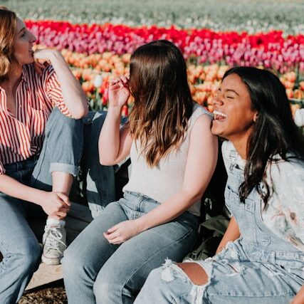 3 women laugh on a park bench with a field of blooming flowers behind them.