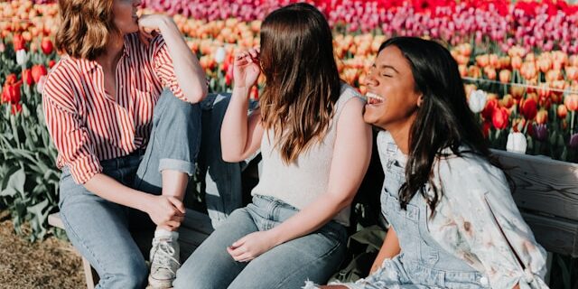3 women laugh on a park bench with a field of blooming flowers behind them.