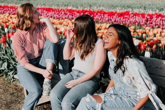 spring-2024-family-and-friends-newsletter 3 women laugh on a park bench with a field of blooming flowers behind them.