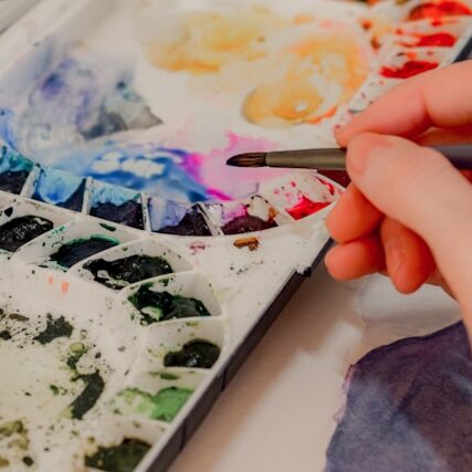 An artist dips their brush in watercolors from a palette.