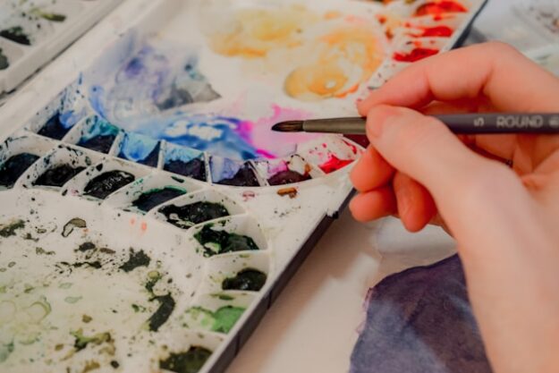 An artist dips their brush in watercolors from a palette.