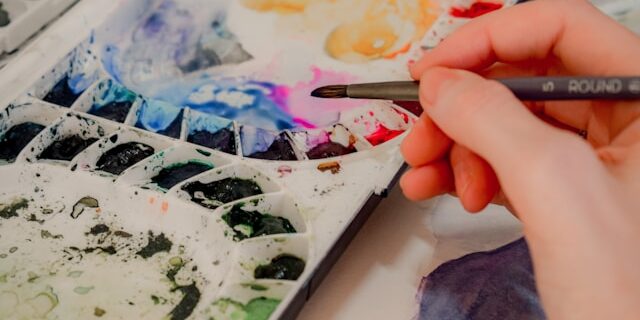 An artist dips their brush in watercolors from a palette.