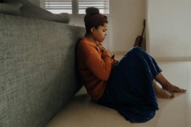 A woman looks stressed as she sits on the floor with her back to a couch.