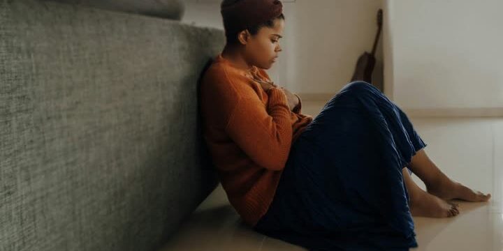 A woman looks stressed as she sits on the floor with her back to a couch.