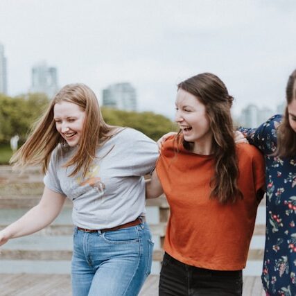 3 friends walk arm-in-arm down a boardwalk near a body of water.