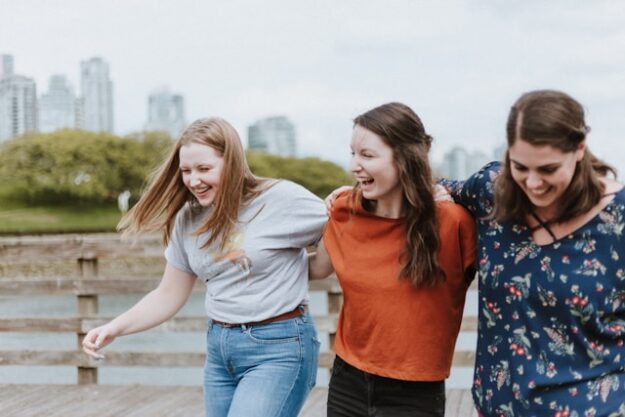 3 friends walk arm-in-arm down a boardwalk near a body of water.