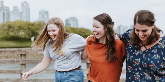 3 friends walk arm-in-arm down a boardwalk near a body of water.