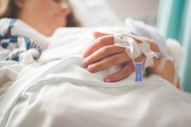 A woman lies in a hospital bed with IV ports taped around her hand.