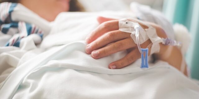 A woman lies in a hospital bed with IV ports taped around her hand.